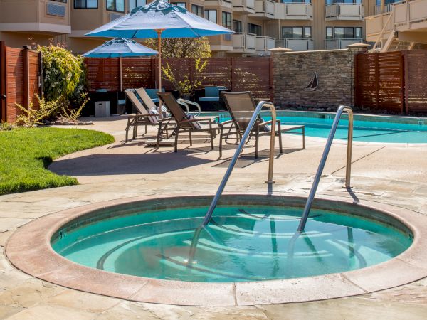 The image shows an outdoor area with a round hot tub and a rectangular swimming pool, surrounded by lounge chairs and umbrellas near a building.