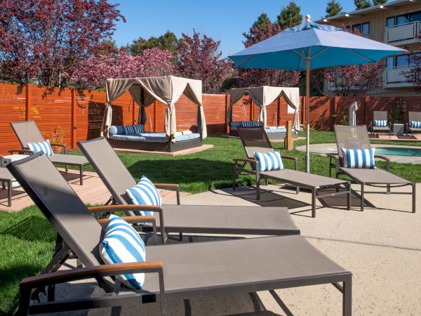 The image shows an outdoor lounge area with sun loungers, striped pillows, canopies, an umbrella, and a pool, surrounded by a wooden fence and trees.