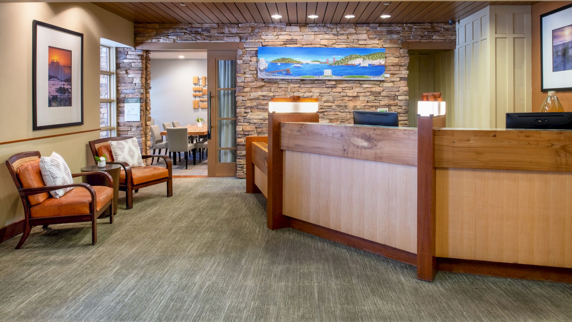The image shows a reception area with wooden counters, chairs with cushions, framed pictures on the walls, and a background meeting area with tables and chairs.