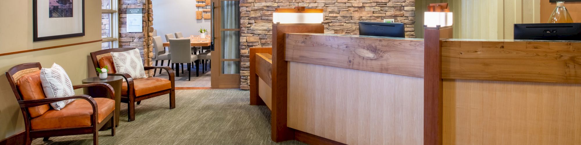 The image shows a reception area with wooden counters, chairs with cushions, framed pictures on the walls, and a background meeting area with tables and chairs.