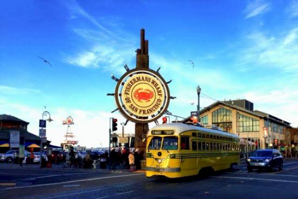 A tram passes by the famous Fisherman's Wharf sign in San Francisco, surrounded by people and buildings under a vibrant blue sky.