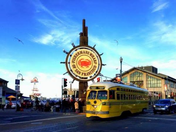 A tram passes by the famous Fisherman's Wharf sign in San Francisco, surrounded by people and buildings under a vibrant blue sky.