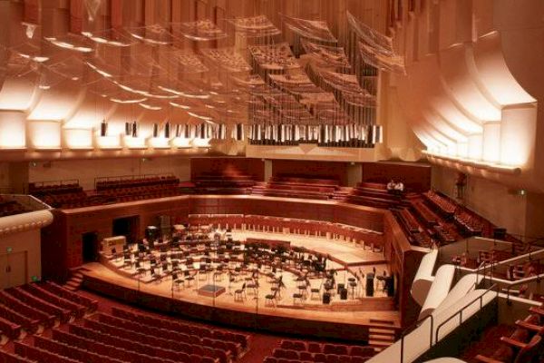 This image shows an empty concert hall with a large stage set up for an orchestra, featuring numerous chairs and music stands under a high ceiling.