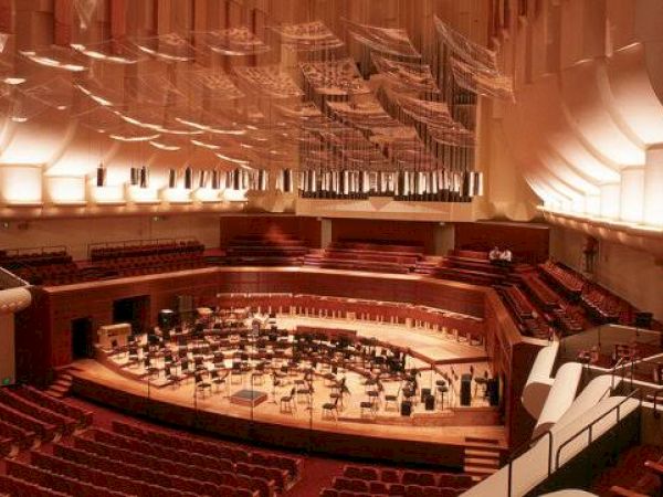 This image shows an empty concert hall with a large stage set up for an orchestra, featuring numerous chairs and music stands under a high ceiling.