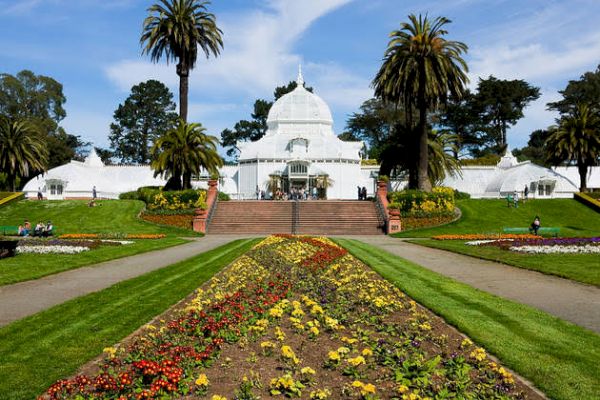 A white conservatory building with a dome, surrounded by colorful flower beds, palm trees, and lush green lawns.