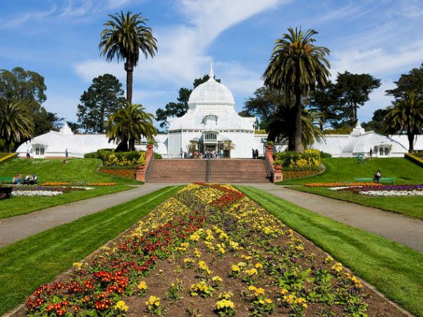 A white conservatory building with a dome, surrounded by colorful flower beds, palm trees, and lush green lawns.