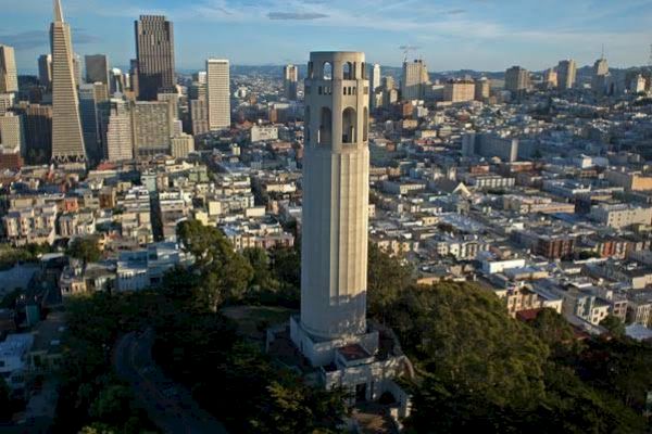 The image shows a tall, cylindrical tower surrounded by a cityscape with numerous buildings and a mix of greenery, likely in an urban area.