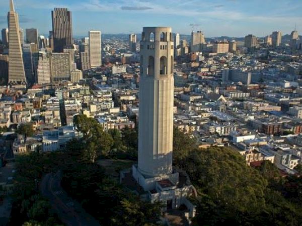 The image shows a tall, cylindrical tower surrounded by a cityscape with numerous buildings and a mix of greenery, likely in an urban area.