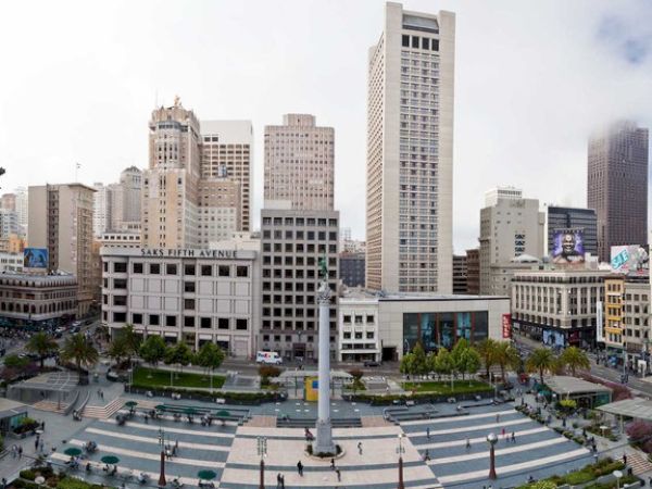 An urban plaza with a tall monument centerpiece is surrounded by high-rise buildings, shops, and trees under a partly cloudy sky.