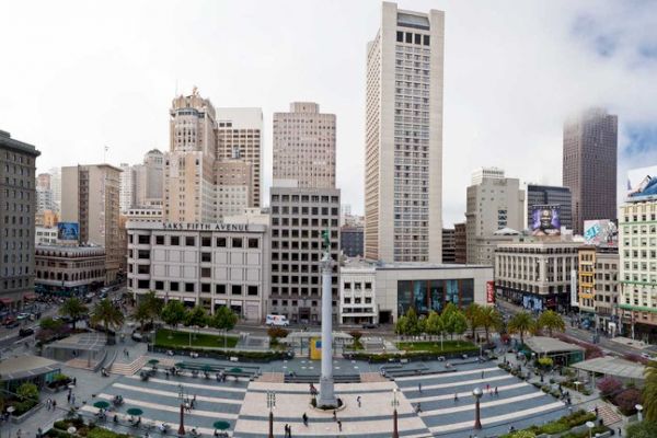 An urban plaza with a tall monument centerpiece is surrounded by high-rise buildings, shops, and trees under a partly cloudy sky.