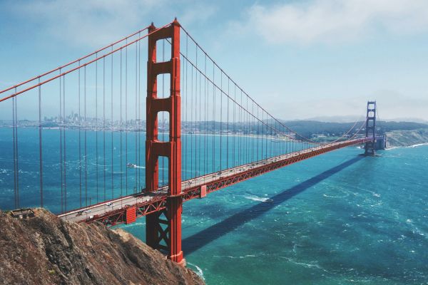The image shows the Golden Gate Bridge, a famous red suspension bridge, spanning a body of water with a cityscape in the background, under a blue sky.