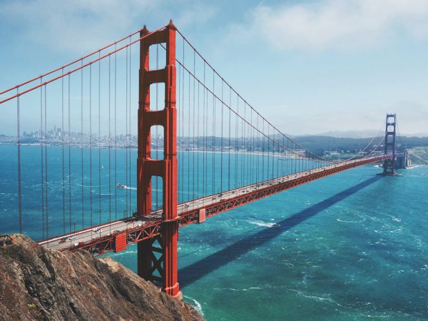 The image shows the Golden Gate Bridge, a famous red suspension bridge, spanning a body of water with a cityscape in the background, under a blue sky.