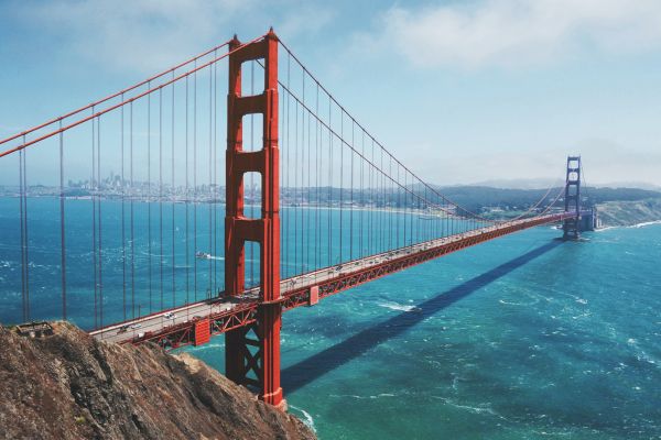 The image shows the Golden Gate Bridge, a famous red suspension bridge, spanning a body of water with a cityscape in the background, under a blue sky.