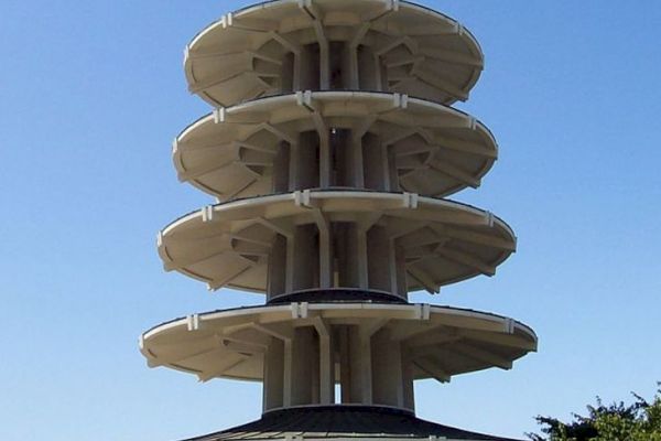 The image shows a modern, multi-tiered pagoda structure against a clear blue sky, surrounded by a few trees and situated in an urban environment.