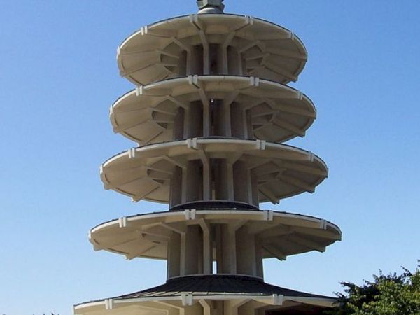 The image shows a modern, multi-tiered pagoda structure against a clear blue sky, surrounded by a few trees and situated in an urban environment.