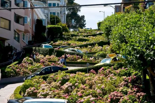 A winding, steep street with tight switchbacks, lined with colorful flowers and greenery, with cars and pedestrians navigating the curves.