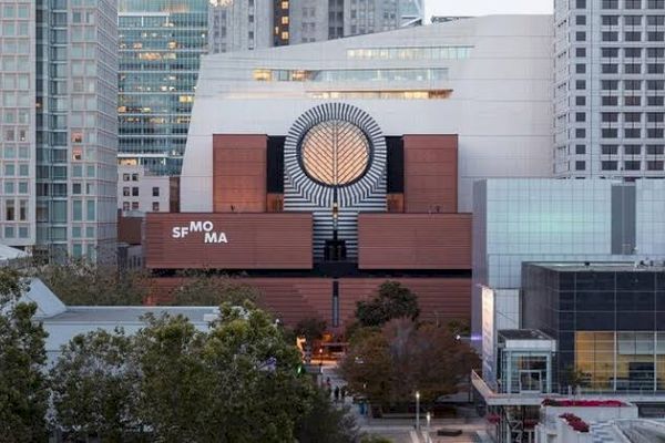 The image shows the San Francisco Museum of Modern Art (SFMOMA) building in an urban setting, surrounded by trees and high-rise buildings.