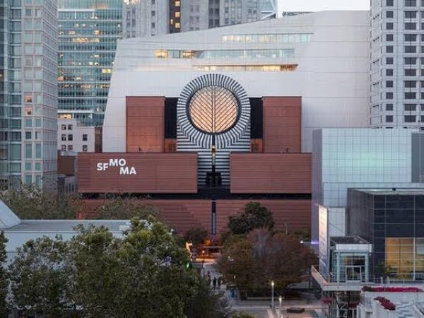 The image shows the San Francisco Museum of Modern Art (SFMOMA) building in an urban setting, surrounded by trees and high-rise buildings.