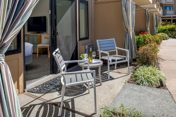 This image shows a cozy outdoor patio area with two chairs and a table, set outside a room with sliding doors, surrounded by plants and flowers.