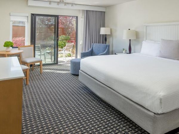 A neatly arranged hotel room with a large bed, TV, armchair, lamp, table with chairs, and a sliding door leading to an outdoor area.