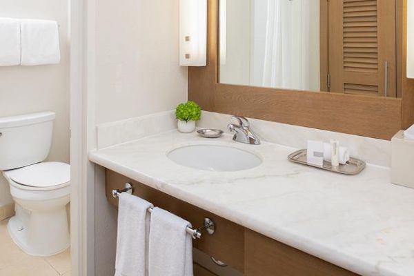 The image shows a modern bathroom with a toilet, a marble countertop sink, a large mirror, and various toiletries organized on the counter.