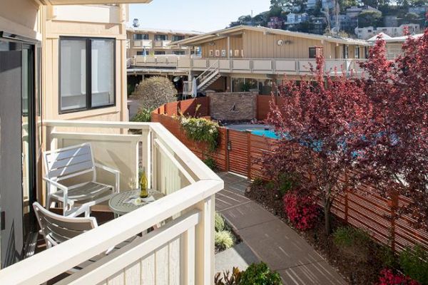 A balcony with chairs and a table overlooks a walkway with red trees and a pool area surrounded by a fence, with buildings and hills in the background.