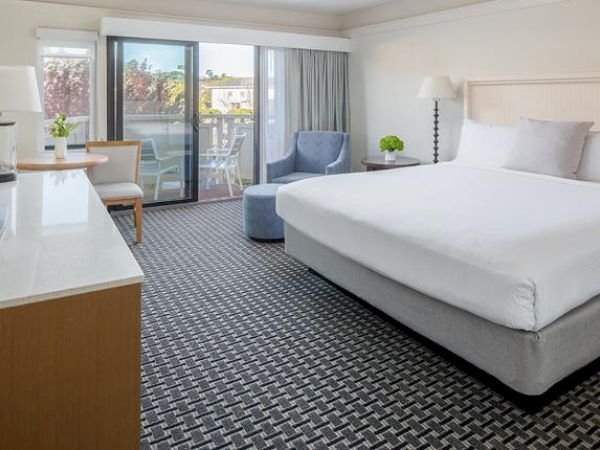 A modern hotel room with a large bed, flat-screen TV, seating area, desk, and a balcony with a view outside, is well-lit and neatly arranged.