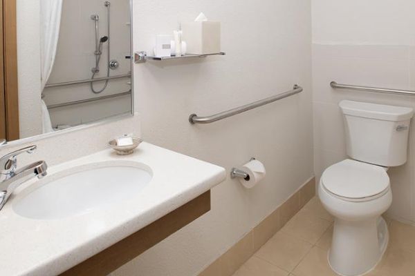 A clean, white bathroom with a sink, toilet, shower, and grab bars for accessibility. There are also toiletries on a shelf above the sink.