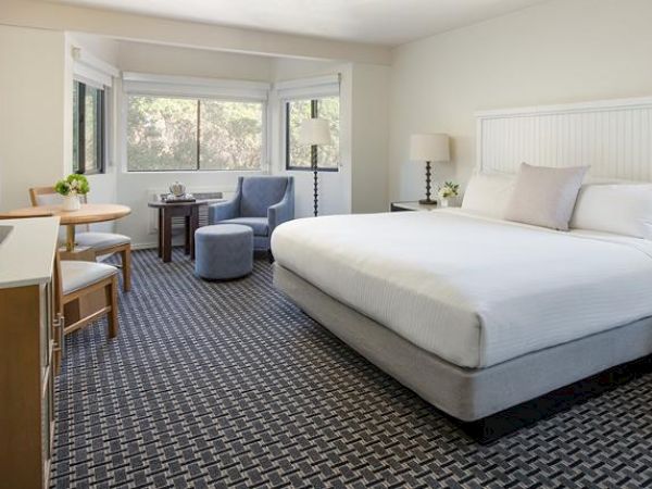 The image shows a hotel room with a king-size bed, a TV, a small dining table with chairs, a lounge chair, and large windows letting in natural light.