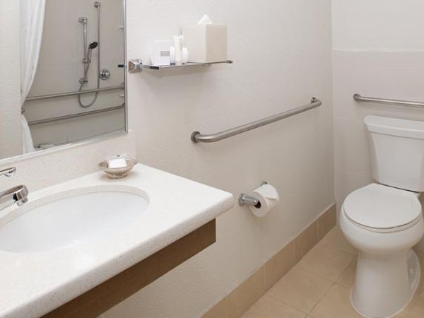 A clean, white bathroom with a sink, toilet, shower, and grab bars for accessibility. There are also toiletries on a shelf above the sink.
