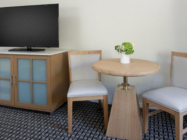 The image shows a small seating area with a round wooden table, two chairs, a cabinet with a TV on top, and a small vase with flowers.
