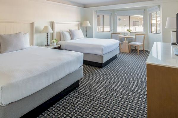 The image shows a hotel room with two double beds, a TV, a small table with two chairs, and large windows, featuring a contemporary and clean design.