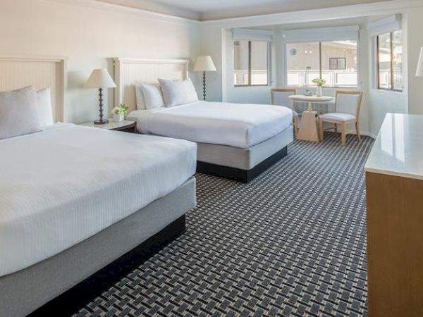 A hotel room with two double beds, a TV, a small table with two chairs, and large windows. The room is bright, with a patterned carpet.