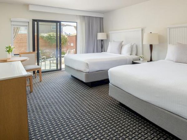 A modern hotel room with two double beds, a flat-screen TV, a desk, and a sliding glass door leading to a balcony or patio.