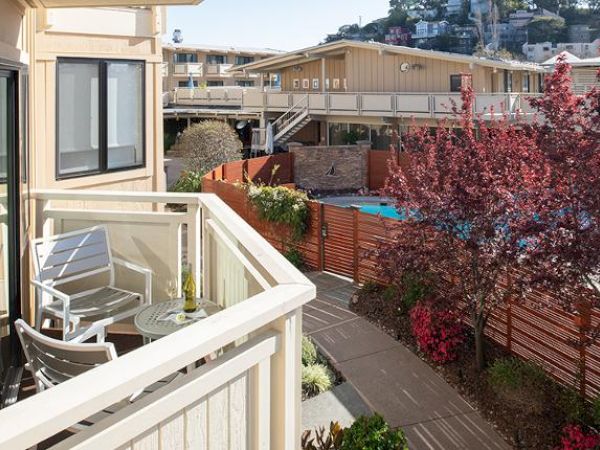 A balcony with chairs and a table overlooks a pool area, surrounded by plants and trees in a residential or hotel setting, with buildings and hills in view.