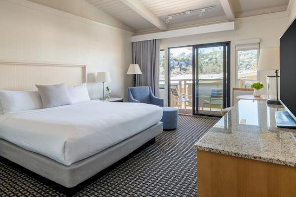 The image shows a modern hotel room with a large bed, an armchair, a TV, and a sliding glass door leading to a balcony with chairs and a table.