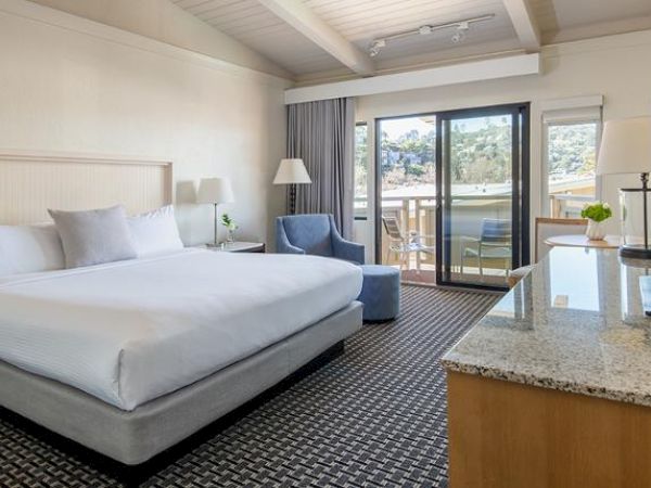 The image shows a modern hotel room with a large bed, an armchair, a TV, and a sliding glass door leading to a balcony with chairs and a table.