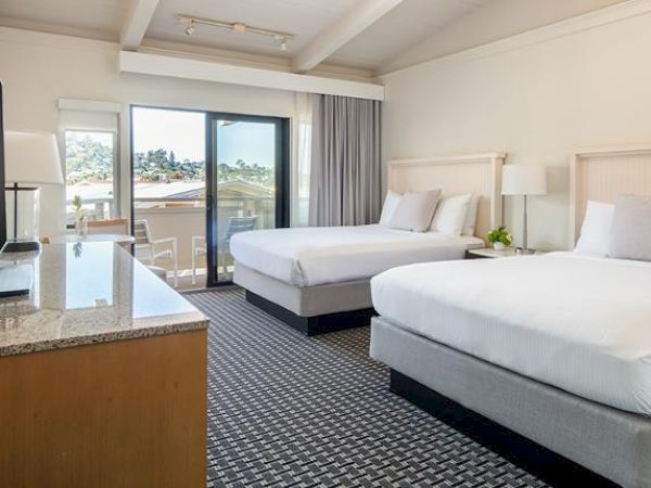 A hotel room with two beds, a TV on a dresser, a window with a view, a small table, and a lamp, all in a bright, neutral-colored setting.