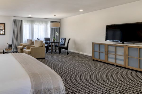 The image shows a spacious hotel room with a large bed, a flat-screen TV, a seating area with chairs and a table, and bright natural light.