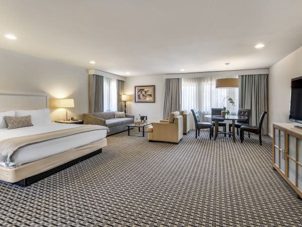 A spacious hotel room features a king bed, sofa, armchairs, table with chairs, flat-screen TV, and large windows, all in neutral tones.
