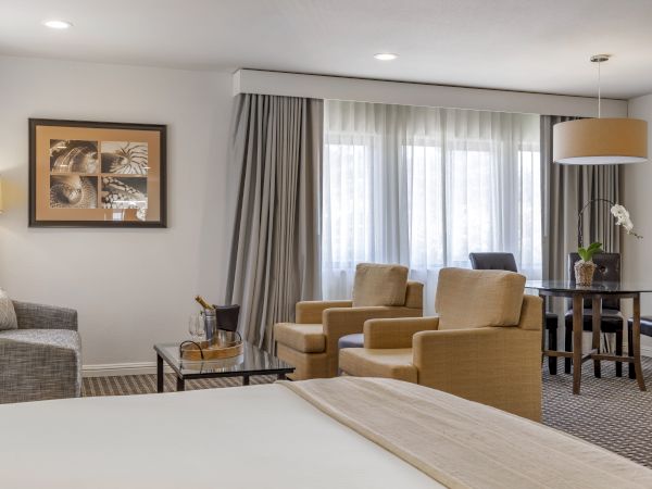 A well-lit hotel room with a bed, sofa, armchairs, table, and lamp. There are large windows with curtains and a framed picture on the wall.