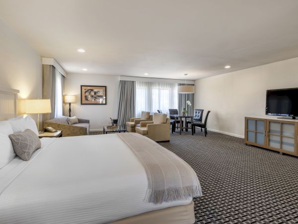 A spacious hotel room featuring a king-sized bed, a seating area with chairs and a sofa, a dining table, a flat-screen TV, and large windows.