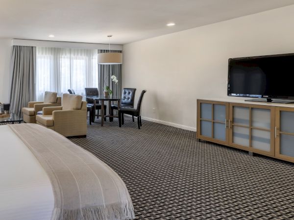 The image shows a spacious hotel room with a large bed, a flat-screen TV, a seating area with chairs and a table, and bright natural light.