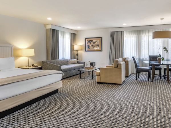 A spacious hotel room features a king-size bed, sofa, armchairs, and a dining table with chairs, all under soft lighting ending the sentence.