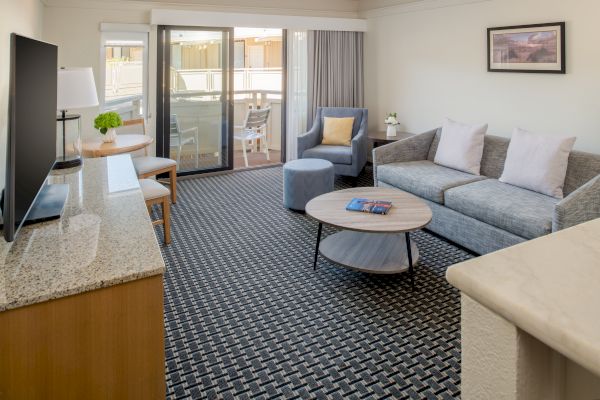 The image shows a modern living room with a sofa, coffee table, TV, and a small balcony. There is a patterned carpet and a piece of art on the wall.