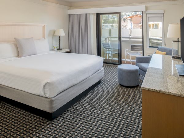 A modern hotel room with a king-size bed, TV, seating area, balcony, and natural light.