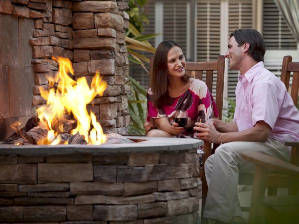 A couple sits on wooden chairs beside a stone fire pit, enjoying drinks and smiling at each other with flames roaring in the background.