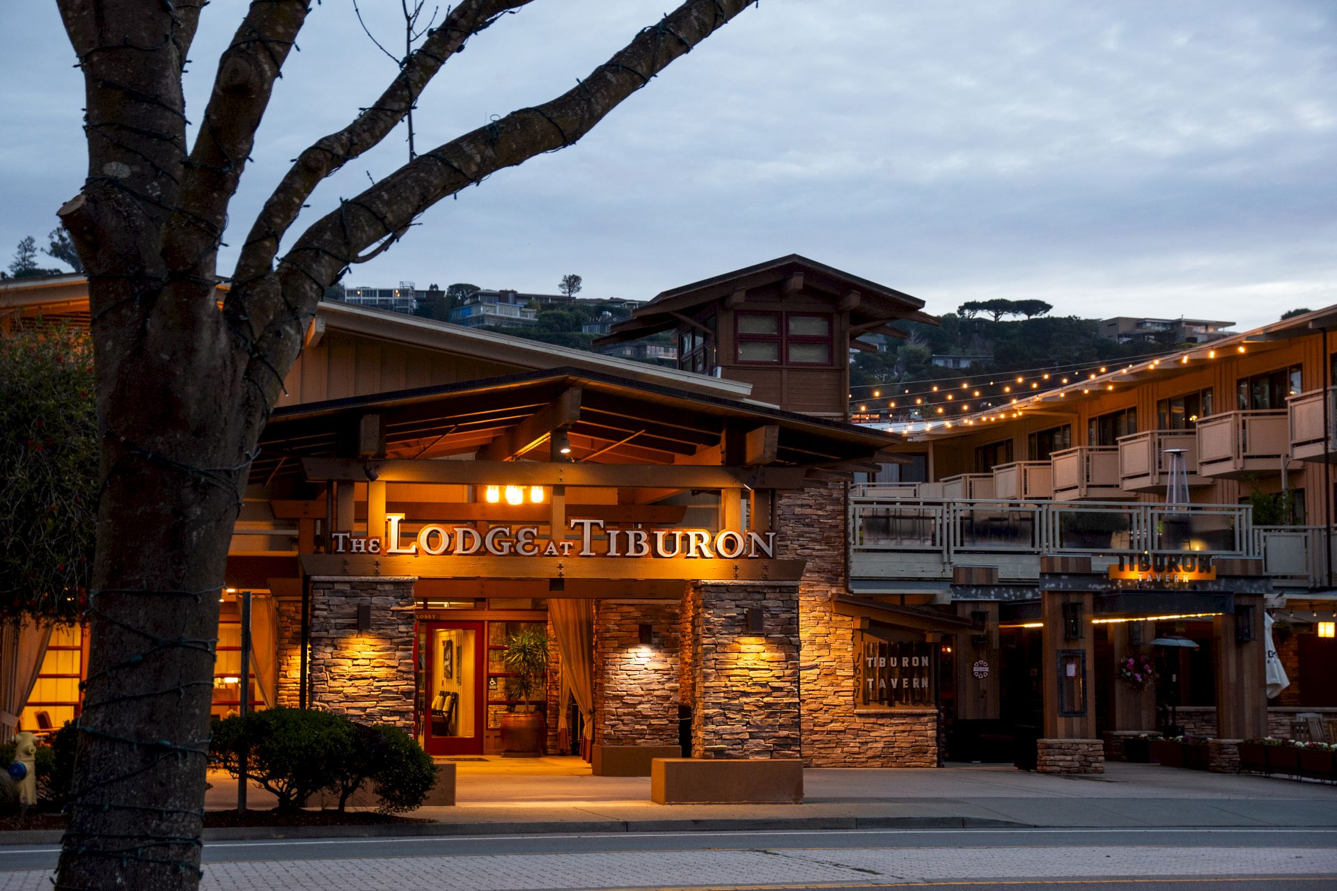 The image shows the exterior of "The Lodge at Tiburon," a building with warm lighting and wooden accents against a background of hills and trees.