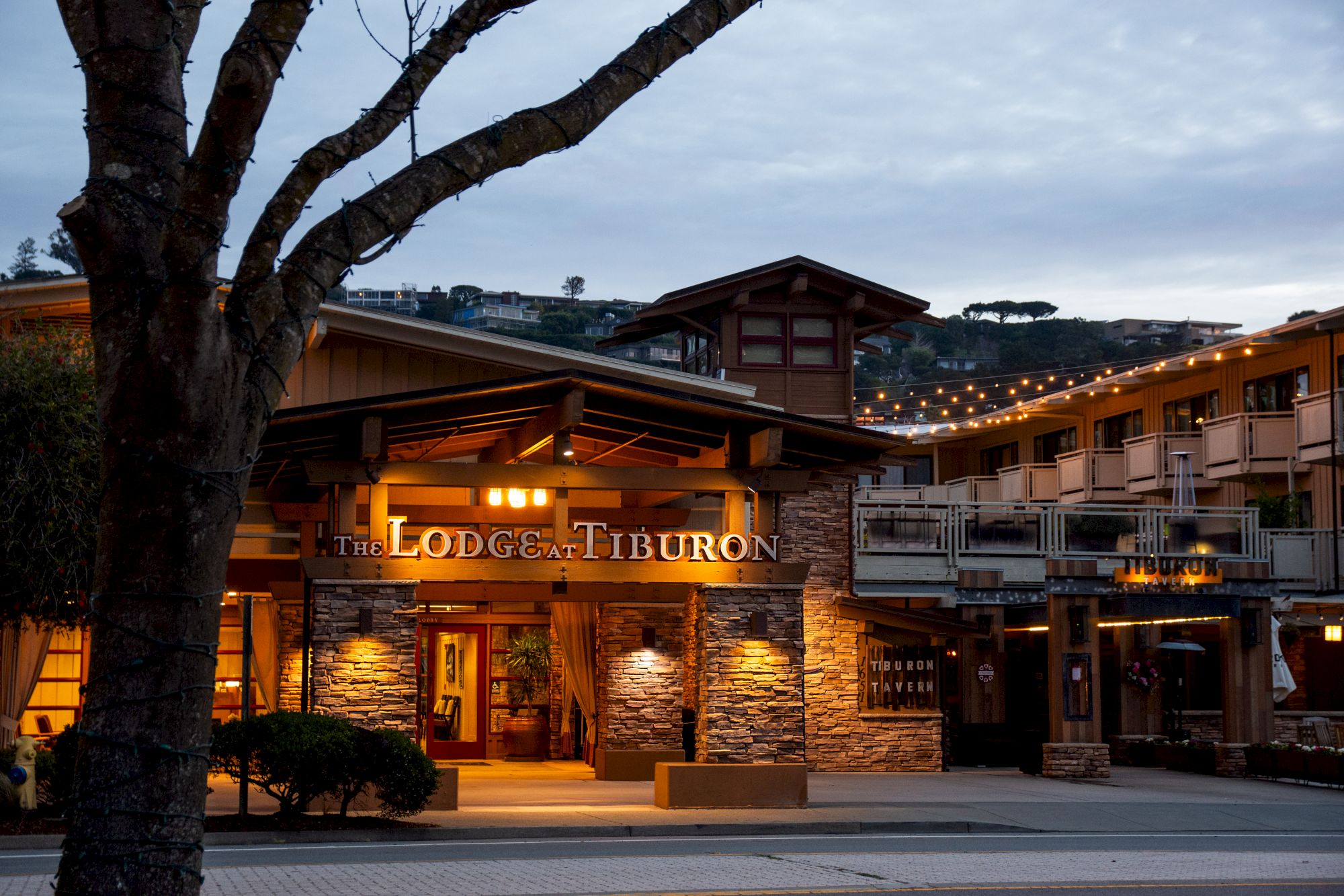 The image shows the exterior of "The Lodge at Tiburon," a building with warm lighting and wooden accents against a background of hills and trees.