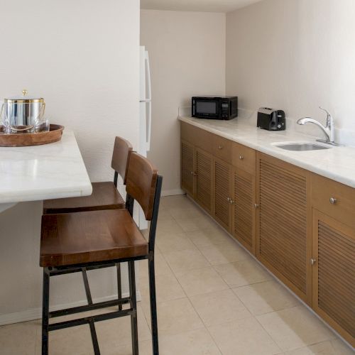 This image shows a narrow kitchen with a countertop, two high chairs, a sink, a microwave, a toaster, a kettle, and wooden cupboards.
